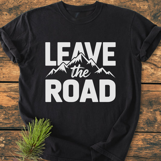 Leave The Road - Vintage Wash Tee, black with bold white slogan and mountain design, laid flat on wood. Pine branch accentuates the garment-dyed, ring-spun cotton's relaxed fit and durability.