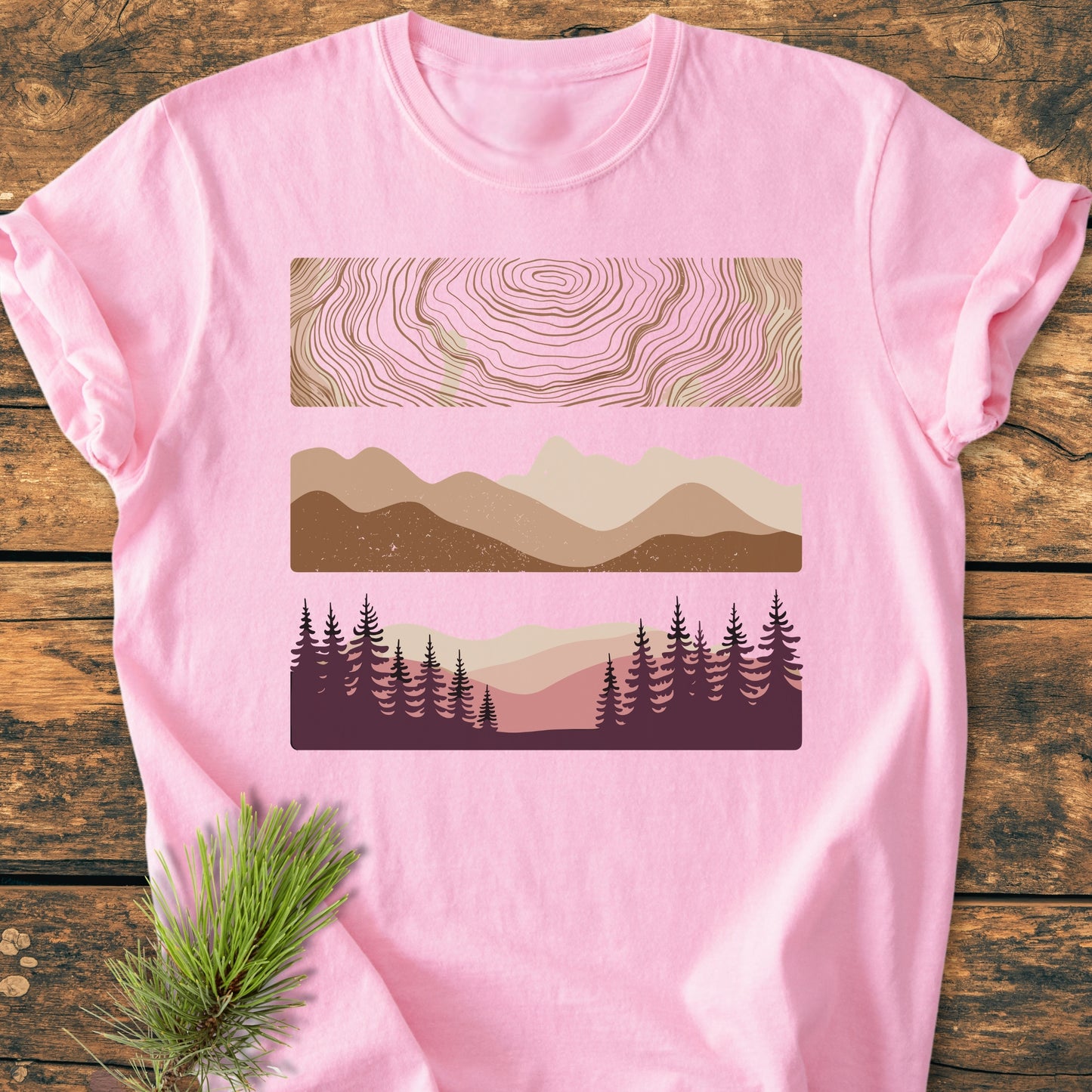 Minimalist Mountain Graphic - Vintage Wash Tee on wood surface, features three graphic panels: contour-line pattern, layered mountains, and a forest scene. Soft pink, made from garment-dyed, pre-shrunk 100% ring-spun cotton.