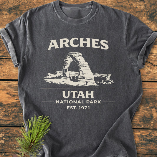 Arches National Park - Vintage Wash Tee features a charcoal-gray hue with ARCHES text and Delicate Arch illustration. Placed on wood, it embodies relaxed comfort and durability with 100% ring-spun cotton.