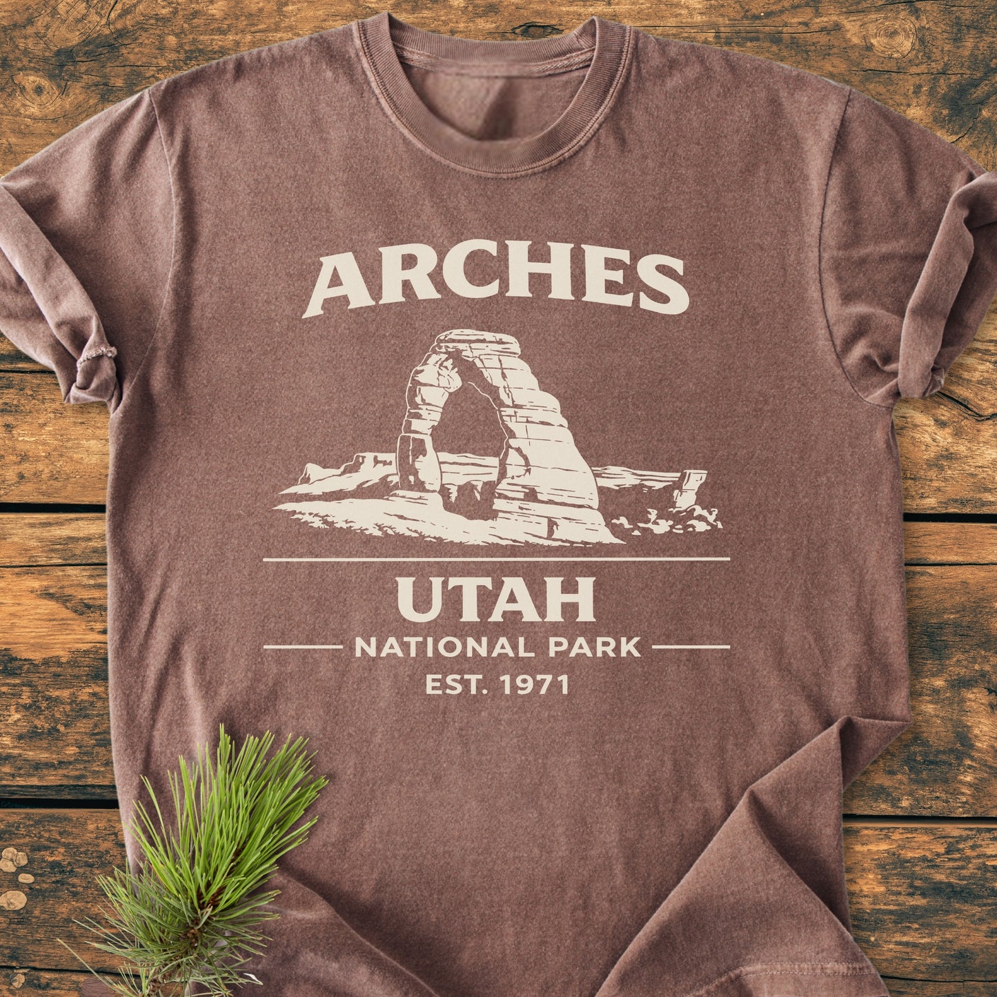 Arches National Park - Vintage Wash Tee folded on wood, features retro print with natural stone arch. Includes soft-washed, garment-dyed fabric, 100% ring-spun cotton, relaxed fit, pine sprig accent.