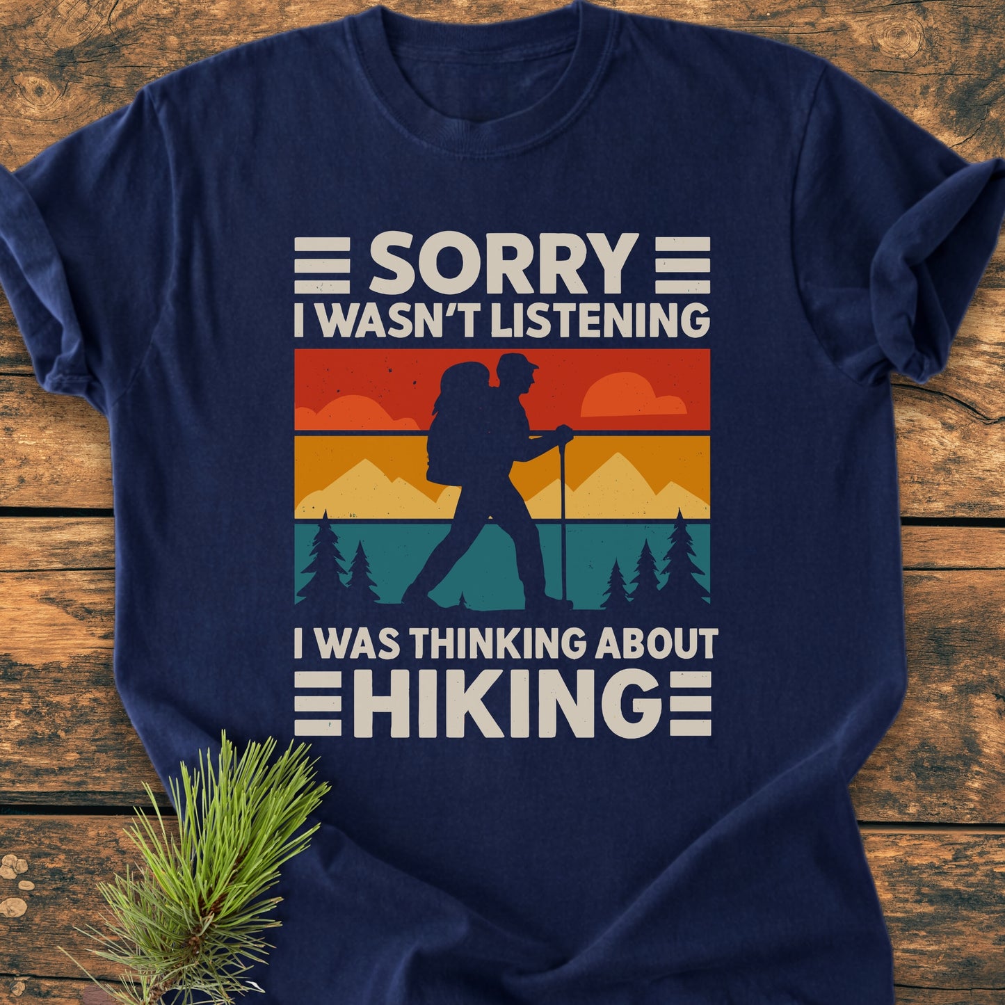 Thinking About Hiking - Vintage Wash Tee: A navy-blue T-shirt featuring a retro graphic of a hiker amidst mountains and sunset, with bold text expressing passion for hiking. Laid on wooden surface with pine sprig.