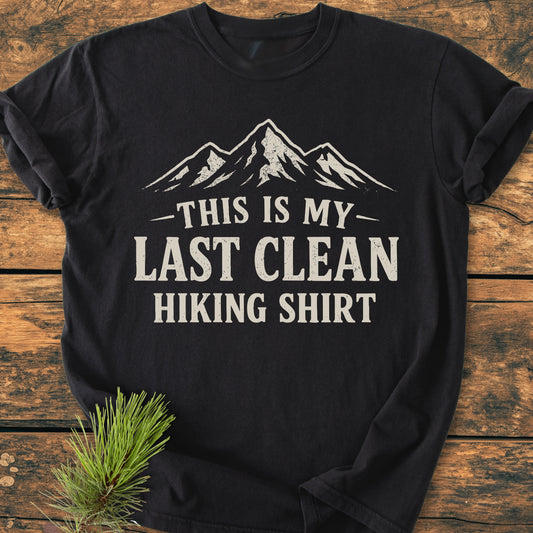 Last Clean Hiking Shirt - Vintage Wash Tee displayed on rustic wood, featuring a mountain graphic and bold text. A pine sprig accents the scene, highlighting its relaxed fit and cozy cotton fabric.