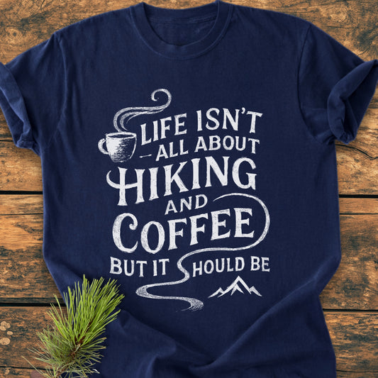 Hiking And Coffee - Vintage Wash Tee features a navy-blue design with a white slogan, coffee-cup icon, and mountain silhouette, emphasizing a rustic, hand-lettered style laid on a wooden surface.