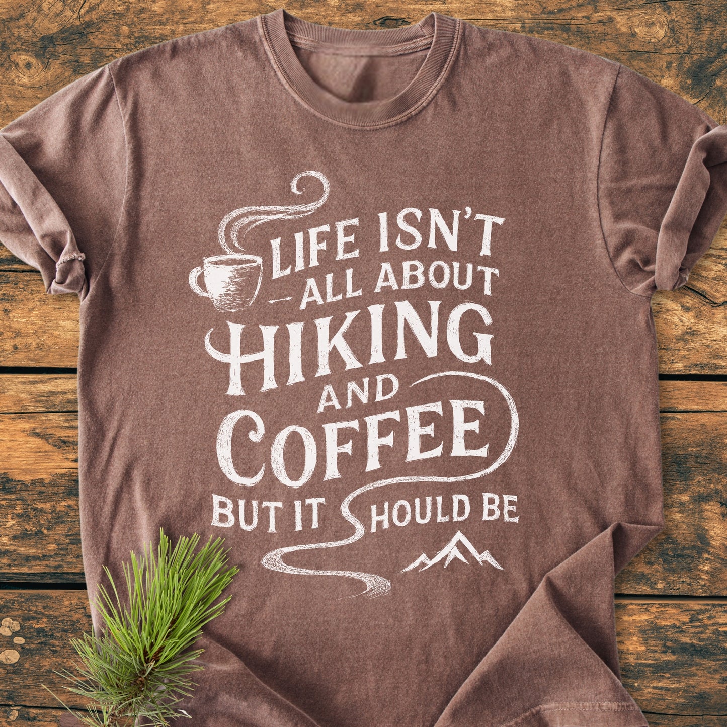 Hiking And Coffee - Vintage Wash Tee: Brown-heathered T-shirt with a white screen-printed quote, coffee-cup icon, and mountain silhouette, laid on wood, accented by a pine branch for an outdoorsy vibe.