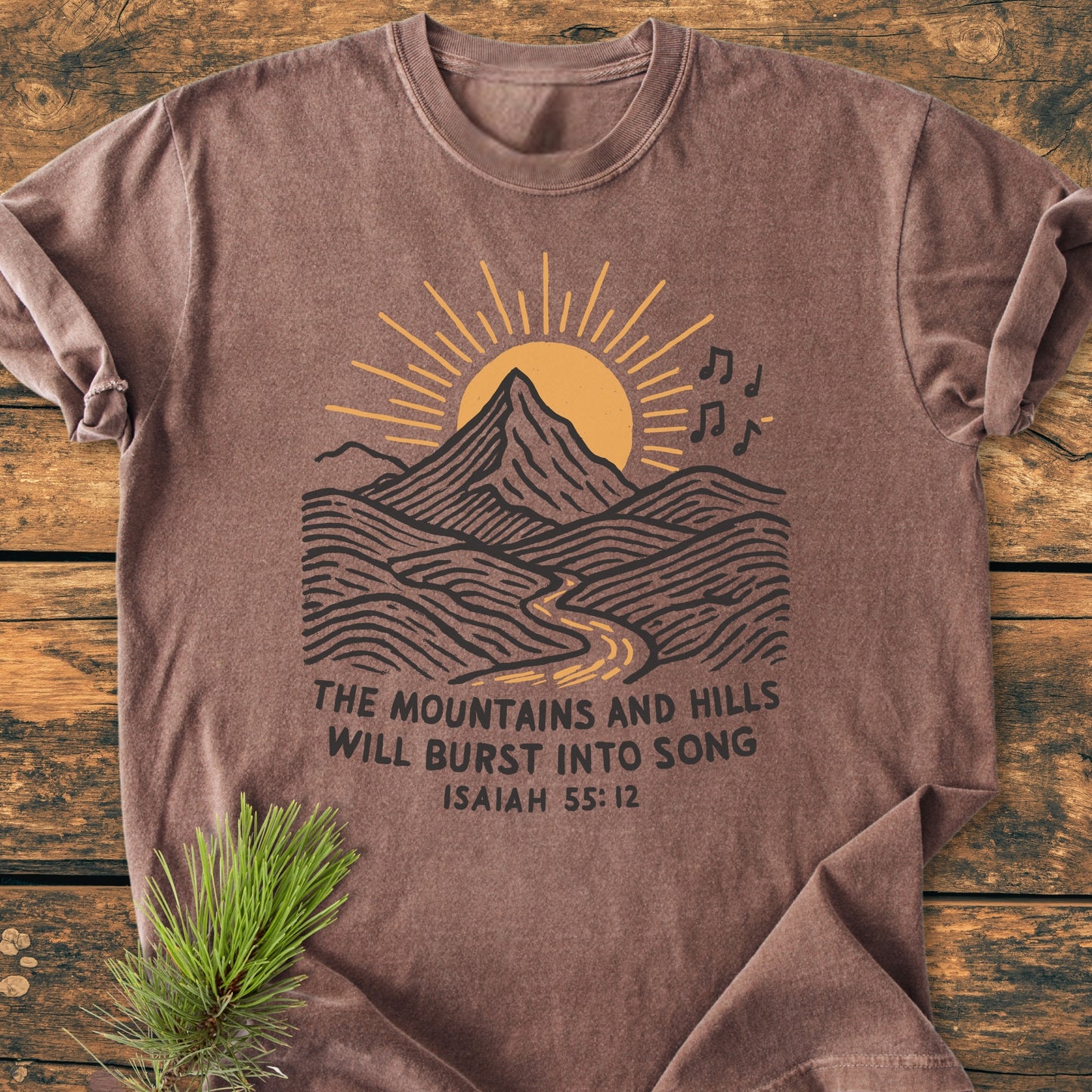Mountains And Hills - Vintage Wash Tee: Brown T-shirt featuring a mountain scene with a rising sun, river, and musical notes, complemented by Isaiah 55:12 text, laid on a wooden surface.