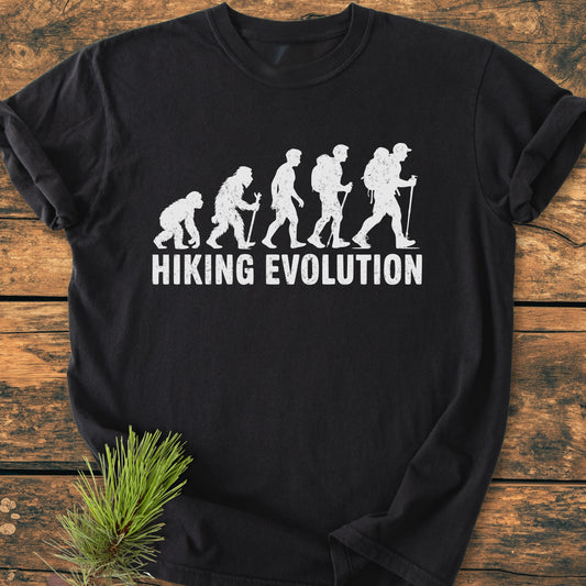 Hiking Evolution - Vintage Wash Tee featuring evolution of man graphic with hiking gear on a black T-shirt, laid on wood. Text HIKING EVOLUTION below. Pine branch accentuates natural theme.