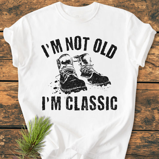 I'm Not Old I'm Classic - Vintage Wash Tee featuring a distressed work boots graphic, laid flat on wood. Decorated with a pine sprig, highlighting its cozy, relaxed fit and durable construction.