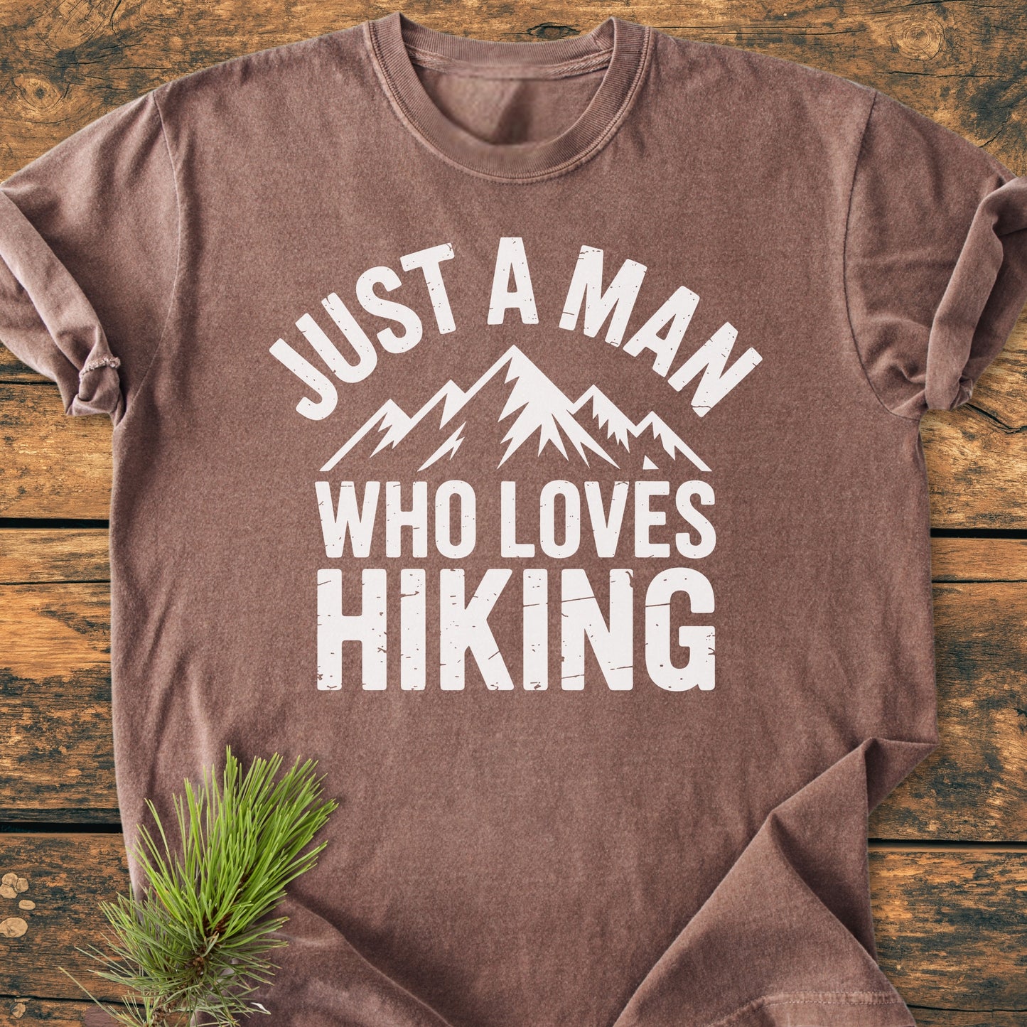 Just A Man Who Loves Hiking - Vintage Wash Tee features white distressed lettering and mountain graphic on a cozy, garment-dyed brown T-shirt, accented with a pine branch on a wooden backdrop.