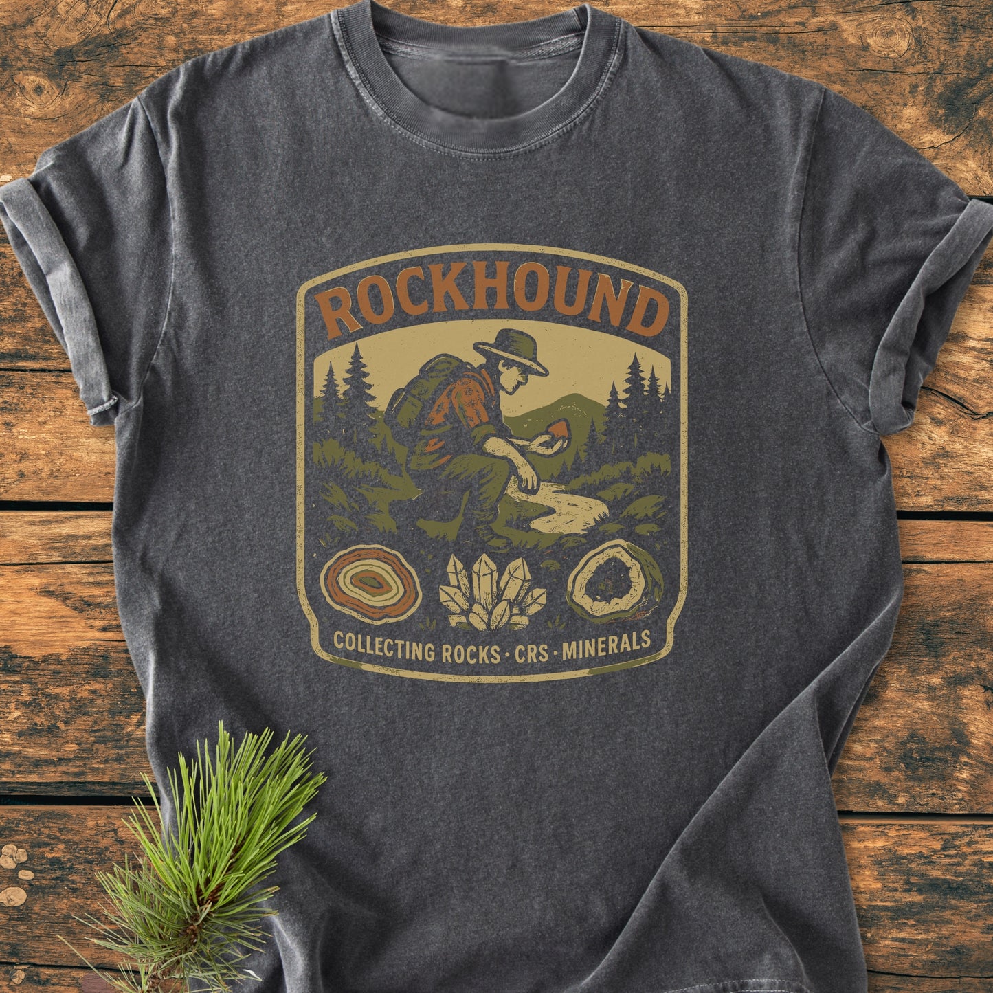 Rockhound Badge - Vintage Wash Tee features a vintage patch graphic of a rock collector in nature, surrounded by mineral illustrations, on a garment-dyed charcoal-gray T-shirt, laid on a wooden surface.
