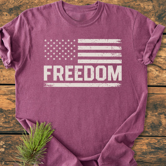 Freedom Flag - Vintage Wash Tee: Heather-purple T-shirt with a distressed white American flag and FREEDOM print, styled with a pine sprig, flat-laid on a wooden surface.