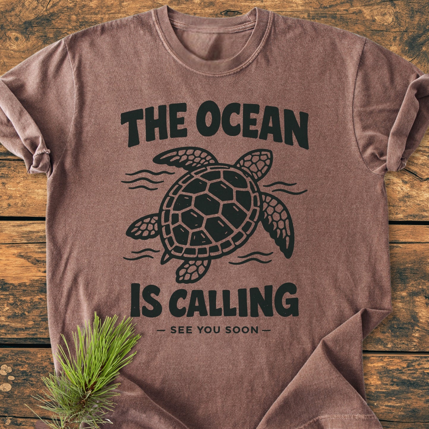 The Ocean Is Calling - Vintage Wash Tee features a sea turtle and slogan design, laid flat with a pine sprig accent, showcasing its crew-neck style and garment-dyed softness.