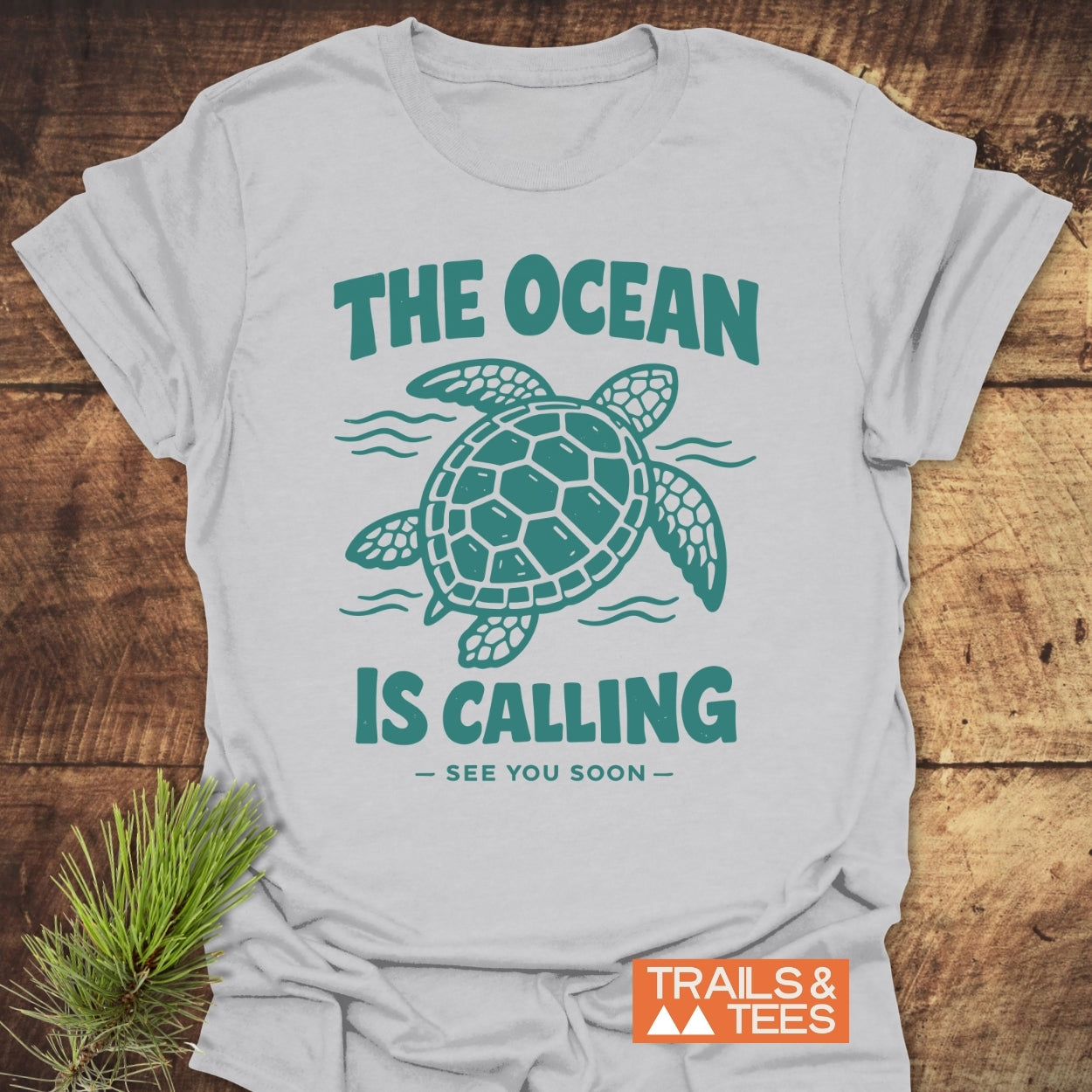 The Ocean Is Calling T-Shirt features a stylized sea turtle graphic with the phrase. Crafted from soft ring-spun cotton, it's designed for comfort and adventure. Trails & Tees logo visible in the corner.
