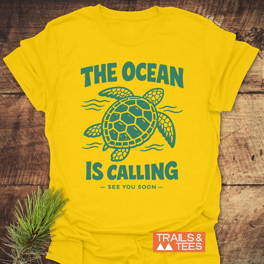 The Ocean Is Calling T-Shirt features a sea turtle and slogan in teal on a yellow crew-neck, laid on wood with a pine sprig. A TRAILS & TEES logo appears on it.