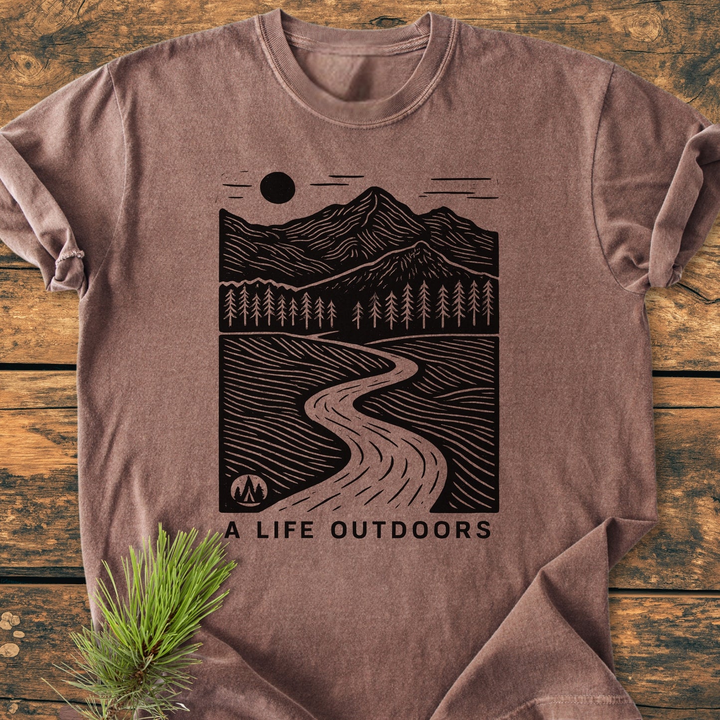 A Life Outdoors - Vintage Wash Tee: Laid-flat earth-tone T-shirt with a black graphic of mountains, pine trees, river, and tent. Slogan “A LIFE OUTDOORS” below. Pine sprig adjacent on wooden background.