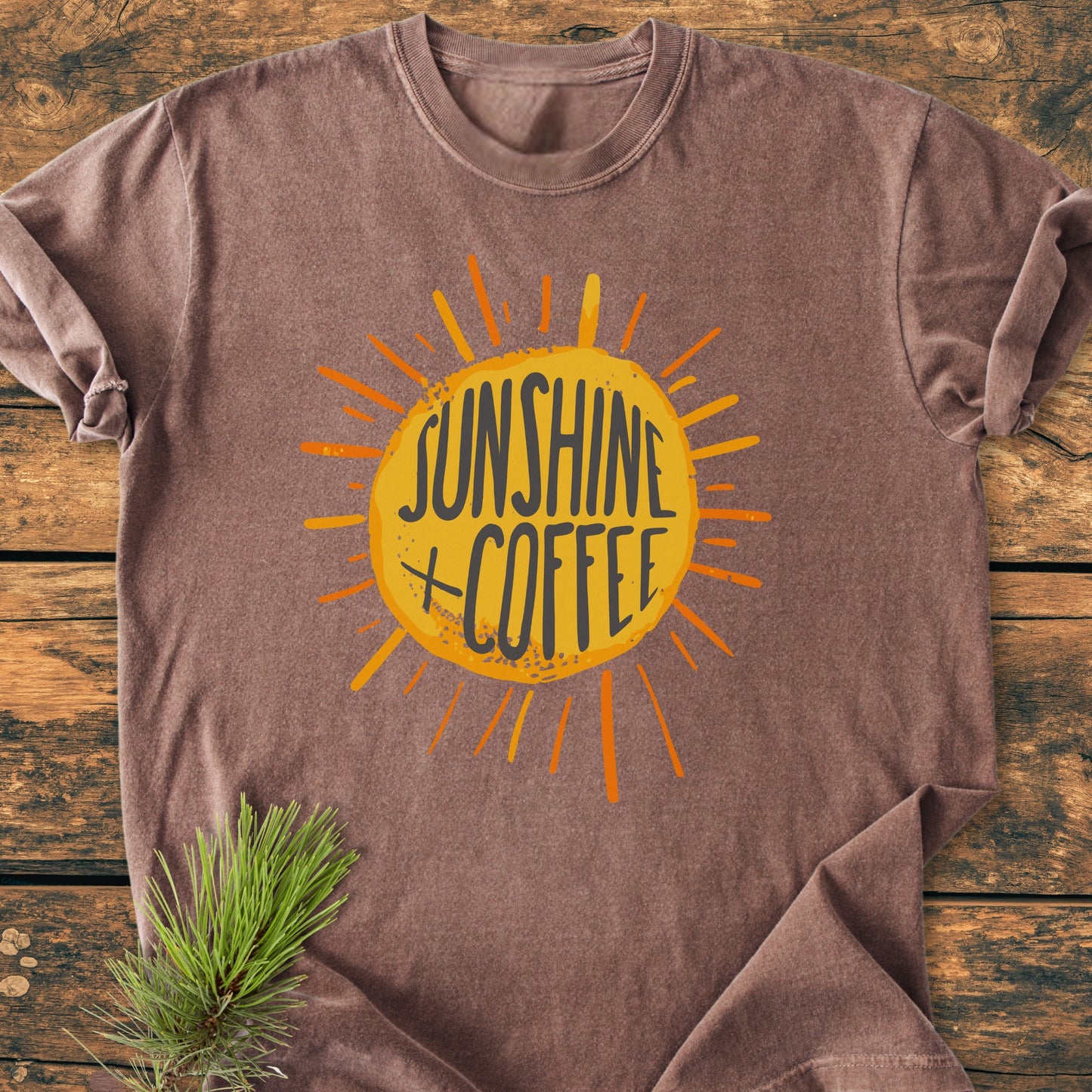 Sunshine + Coffee - Vintage Wash Tee: A brown short-sleeve T-shirt featuring a yellow sun graphic with SUNSHINE COFFEE text, laid flat on a wooden surface with a small pine branch nearby.
