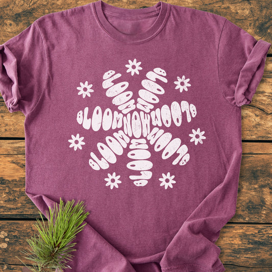 Bloom Now - Vintage Wash Tee in heathered purple, featuring a central white stylized snowflake graphic, displayed flat on wood with a pine sprig, emphasizing its cozy, relaxed fit and durable design.