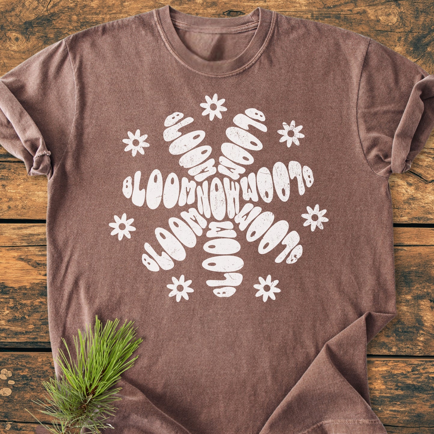 Bloom Now - Vintage Wash Tee: A brownish-mauve T-shirt with a white bloom now snowflake motif, displayed on a wooden surface, accented by a pine sprig at the edge.