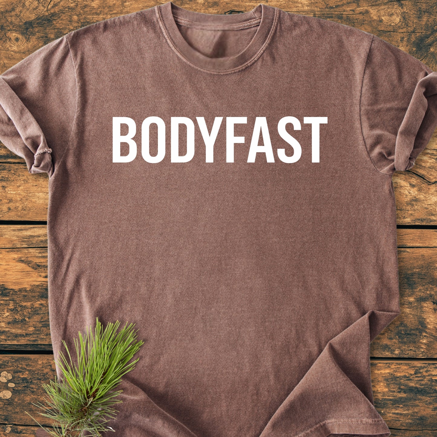 BODYFAST - Vintage Wash Tee features prominently in white letters on a brown t-shirt, laid on wood, with a pine sprig accentuating the rustic, lifestyle-inspired design.