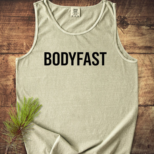 Light-heather gray BODYFAST Tank Top, sleeveless, on rustic wood, features bold black lettering. Beside it, a pine sprig adds a natural touch, highlighting the garment's relaxed, sustainable design.
