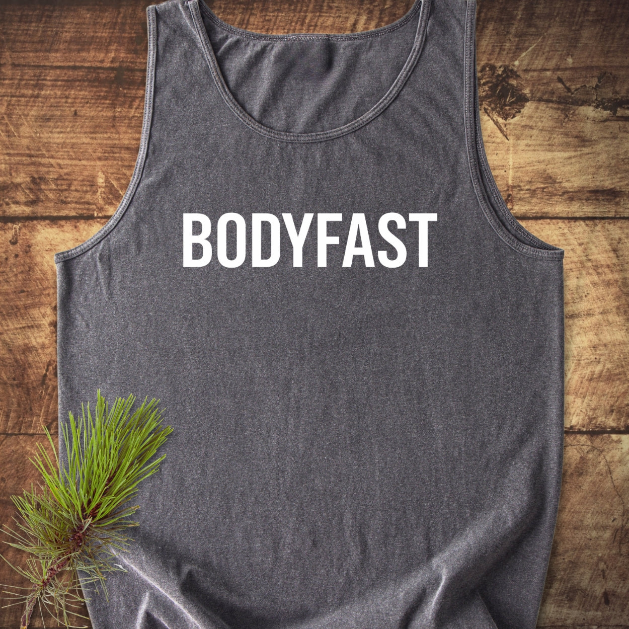BODYFAST Tank Top: Charcoal-gray muscle shirt with bold white 'BODYFAST' text, laid flat on a wooden surface, featuring a small green pine-needle sprig accent, highlighting eco-friendly and stylish design.
