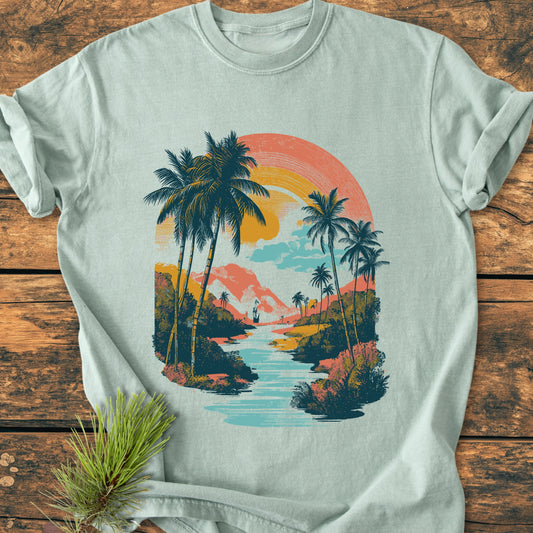 Along The River - Vintage Wash Tee features a retro tropical scene with a river, palm trees, and a setting sun in muted pastels, laid on a rustic surface.