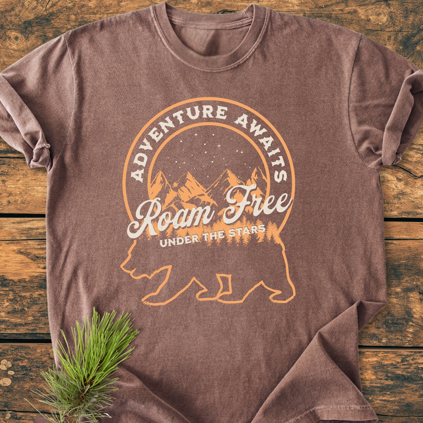 Roam Free Bear - Vintage Wash Tee with stylized bear and outdoor scene, laid flat on wood, sleeves rolled; soft, durable, pre-shrunk 100% cotton, relaxed fit, ideal for adventure lovers.