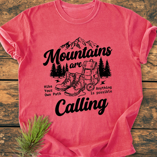 Cute Mountains Are Calling Illustration - Vintage Wash Tee features a mountain range, trees, hiking gear, and motivational slogans. Laid flat on wood with pine needles accent, showcasing its cozy, relaxed fit.