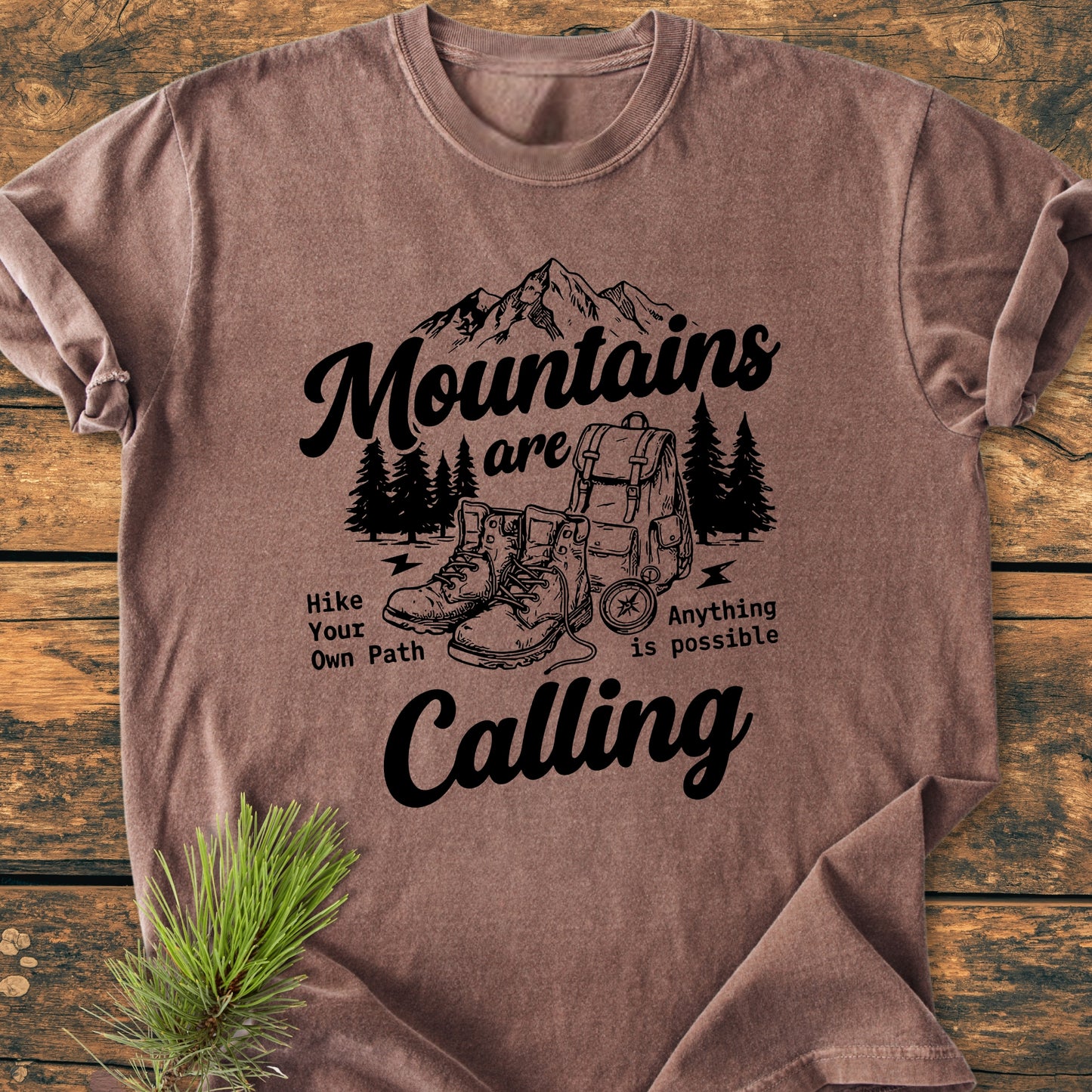 Cute Mountains Are Calling Illustration - Vintage Wash Tee featuring a black outdoor-adventure graphic with mountains, pine trees, hiking gear, and motivational text, laid flat on wood with a pine sprig accent.