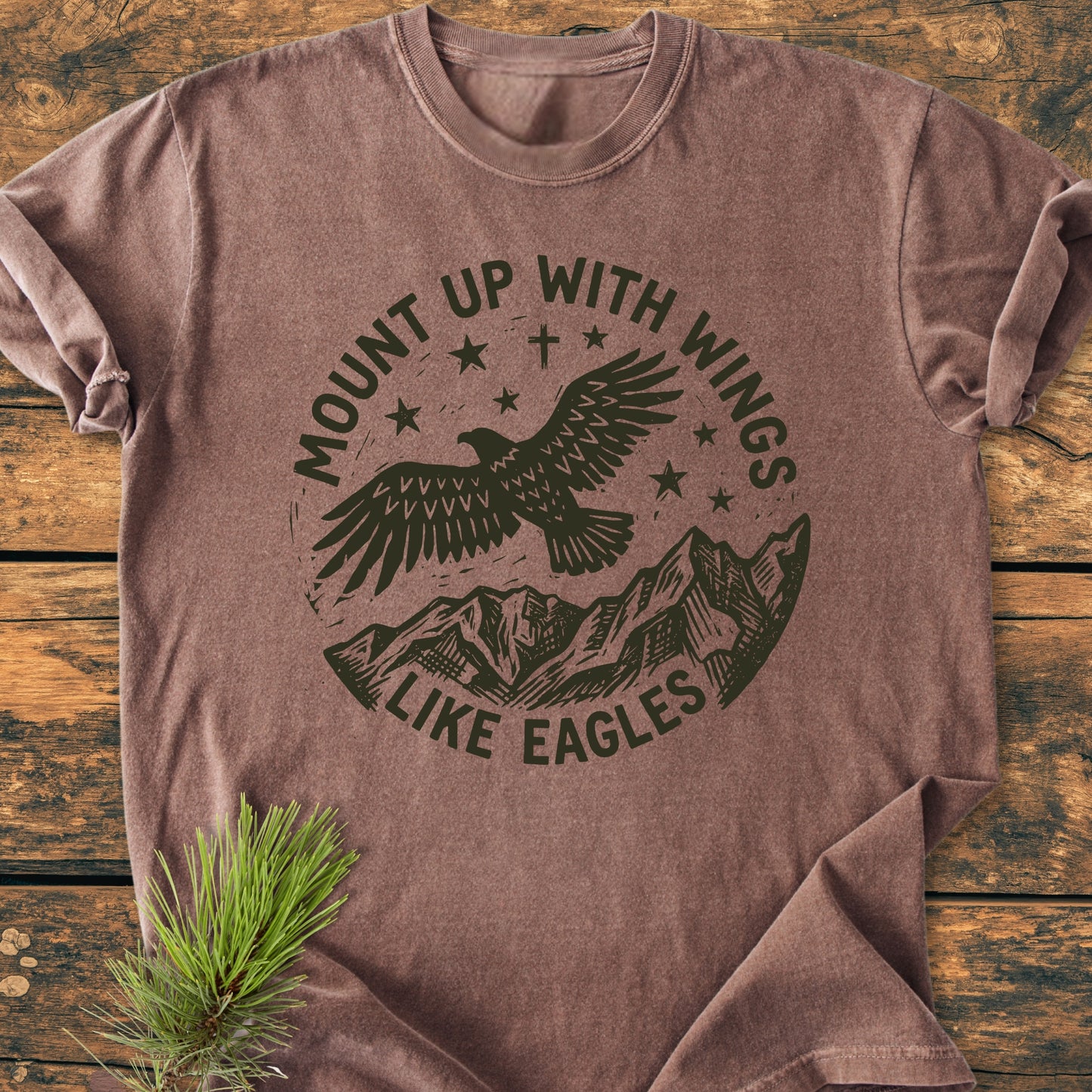 Mount Up With Wings - Vintage Wash Tee featuring a hand-drawn eagle and mountain graphic on a brown-toned shirt, laid on wood. Displays MOUNT UP WITH WINGS and LIKE EAGLES slogans.