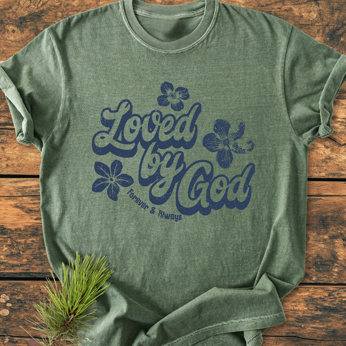 Loved By God - Vintage Wash Tee features a navy hand-lettered design with floral accents on a green-heather backdrop, laid on rustic wood, with an evergreen sprig accentuating the scene.