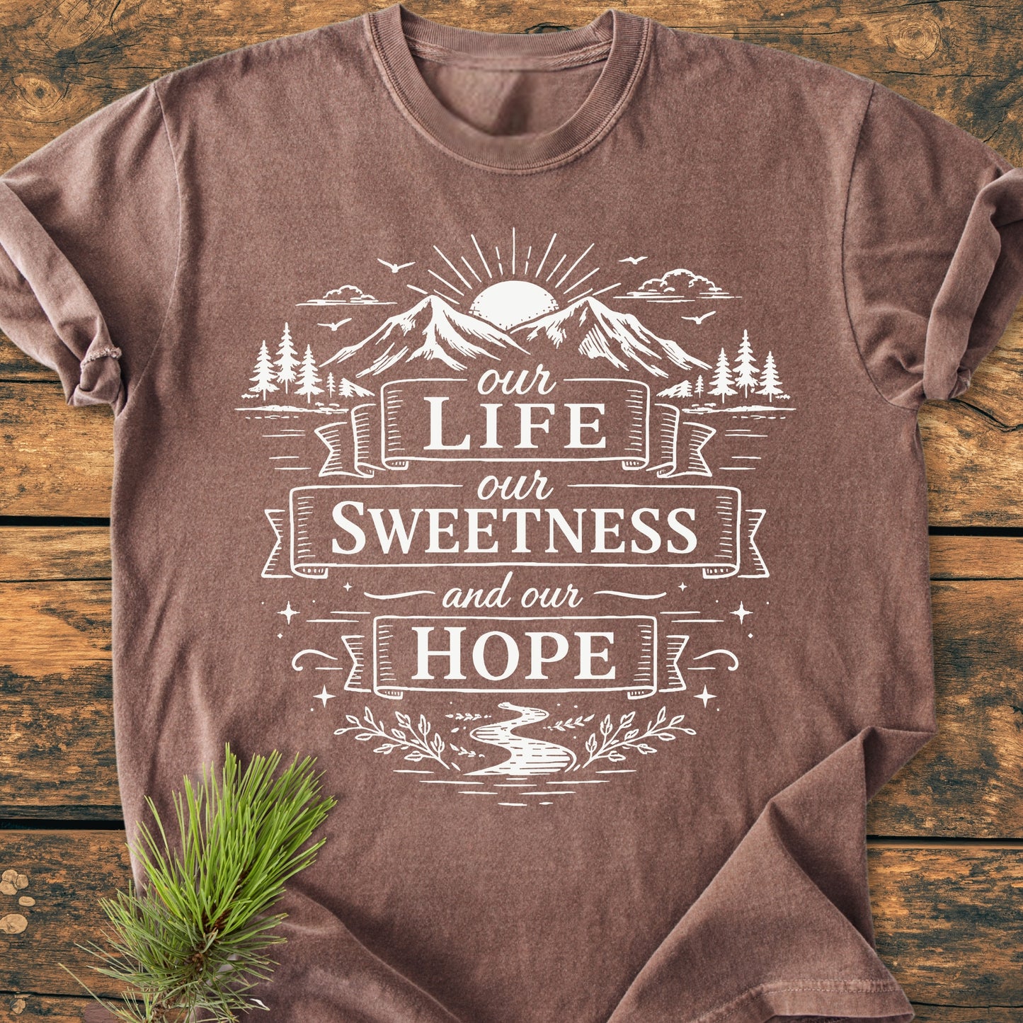 Our Life, Our Sweetness, Our Hope - Vintage Wash Tee features a nature-inspired design with a sun, mountains, pine trees, and a river motif, printed on a cozy, brown short-sleeve T-shirt.