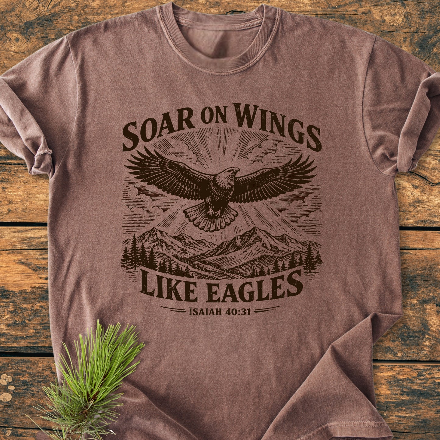 Soar On Wings Like Eagles Isaiah 40:31 - Vintage Wash Tee featuring an eagle graphic over mountains with sun-rays, displayed on a rustic surface with a pine sprig accentuating the scene.