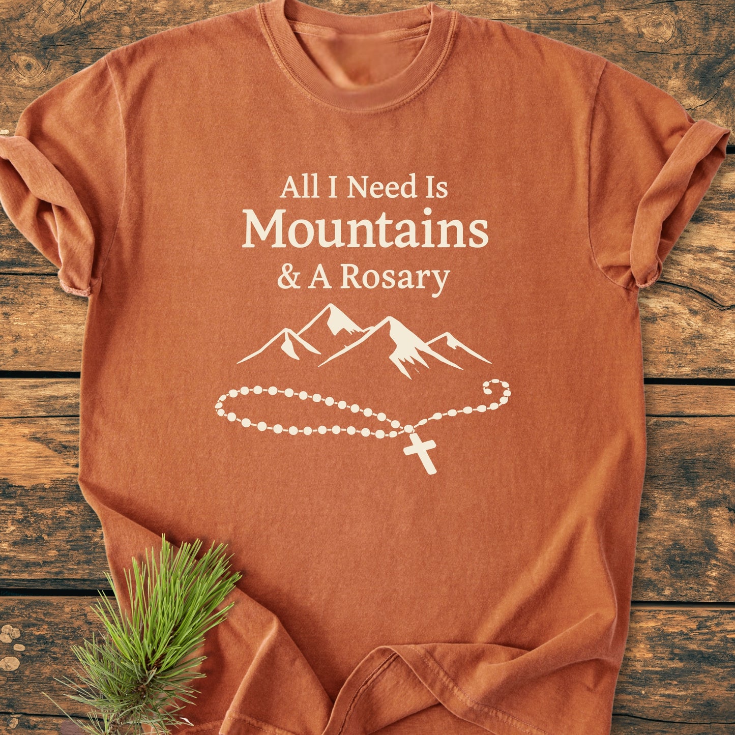 The All I Need Is Mountains And A Rosary - Vintage Wash Tee features a rust-orange hue with white text and graphics, styled with pine on a wooden backdrop, highlighting its rustic, outdoor-inspired design.