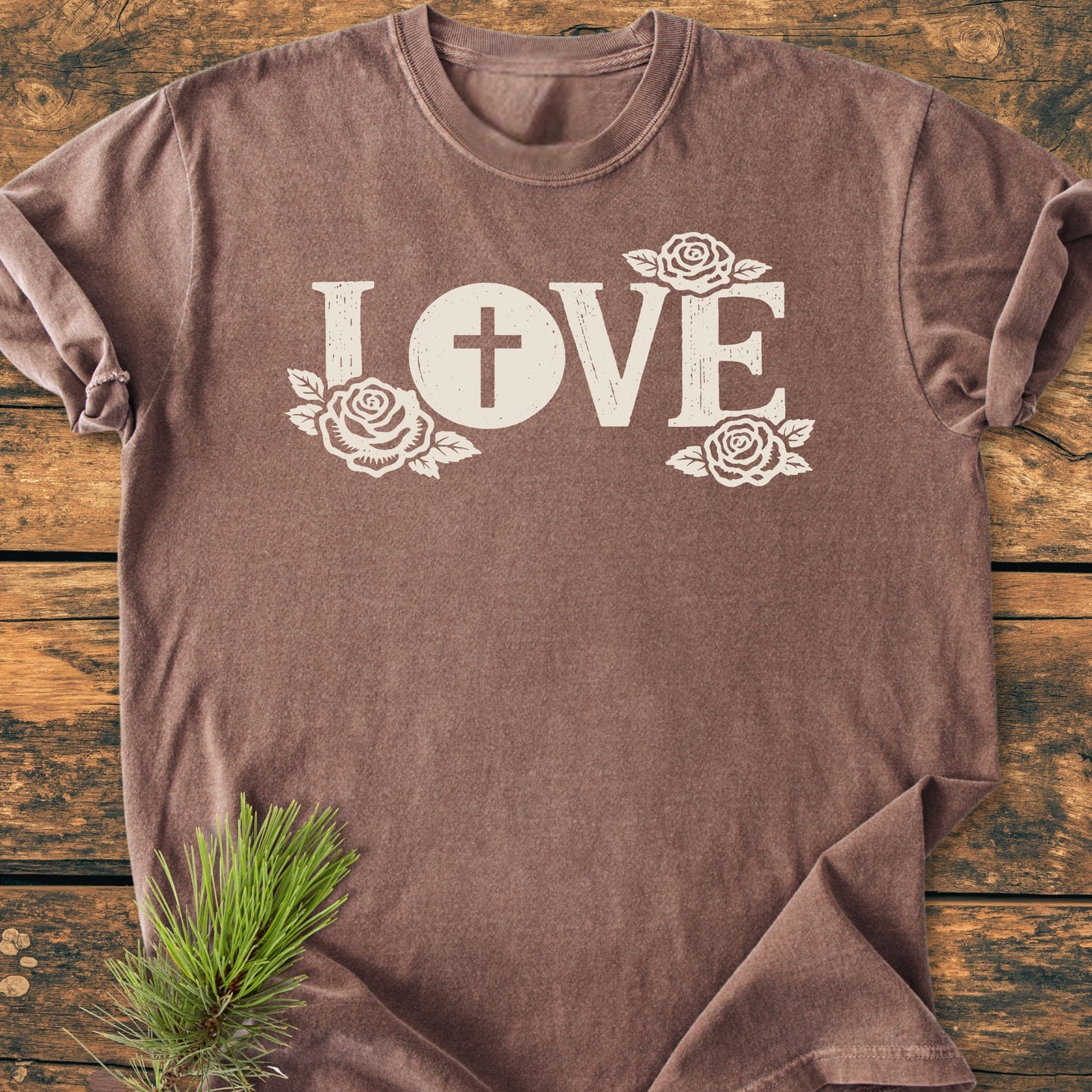 LOVE – Eucharistic Faith - Vintage Wash Tee: A brown, short-sleeved T-shirt featuring LOVE with a cross in the O and rose illustrations, laid on wood with pine needles.