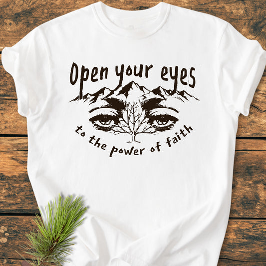 Open Your Eyes To The Power Of Faith - Vintage Wash Tee featuring a mountain and eyes graphic on soft, garment-dyed cotton, displayed with a pine sprig on a wooden surface.