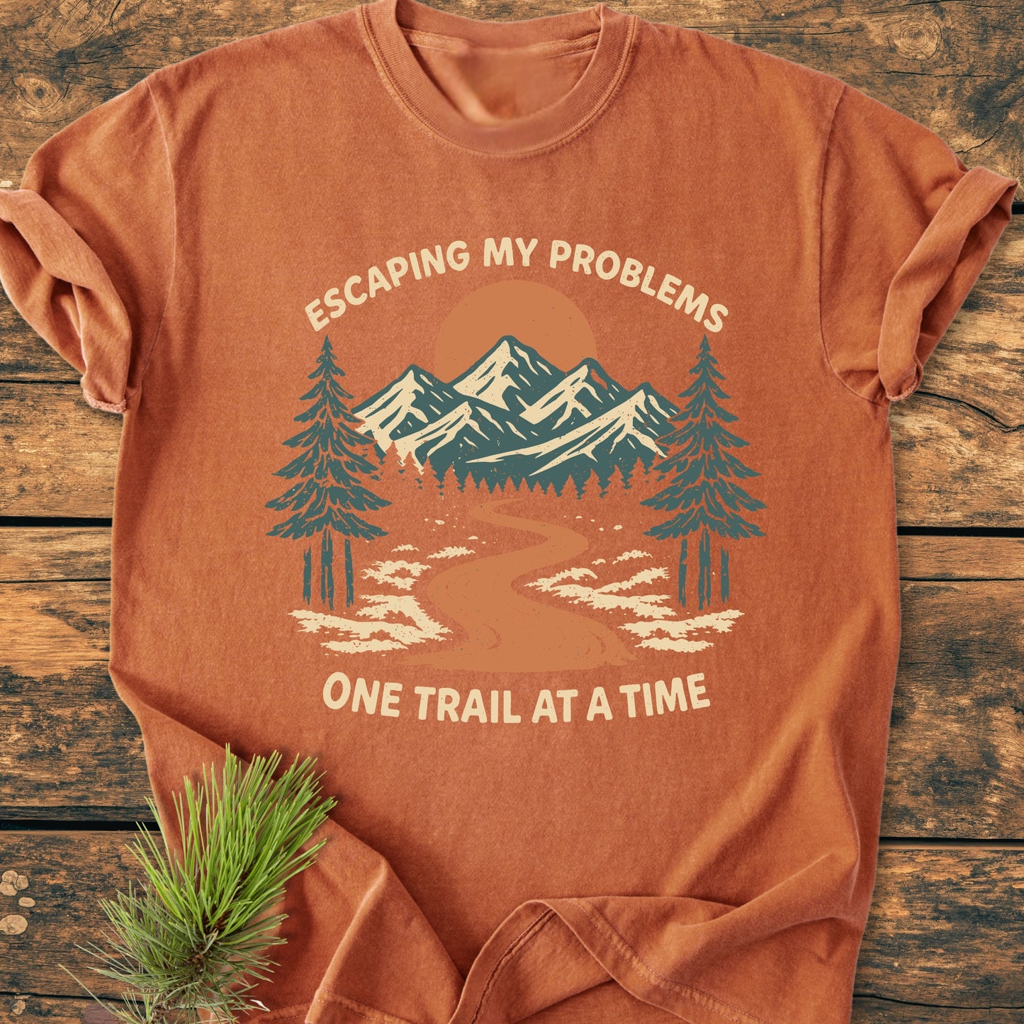 Escaping My Problems - Vintage Wash Tee: Rust-orange T-shirt with nature graphic of pine trees, trail, mountains, and sun, adorned with 'ESCAPING MY PROBLEMS, ONE TRAIL AT A TIME.' Pine sprig beside.