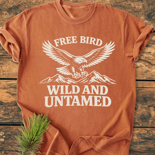 Free Bird - Vintage Wash Tee featuring a stylized eagle and mountains print, laid flat on a wooden surface with a pine sprig beside it. Cozy, durable, and relaxed fit.