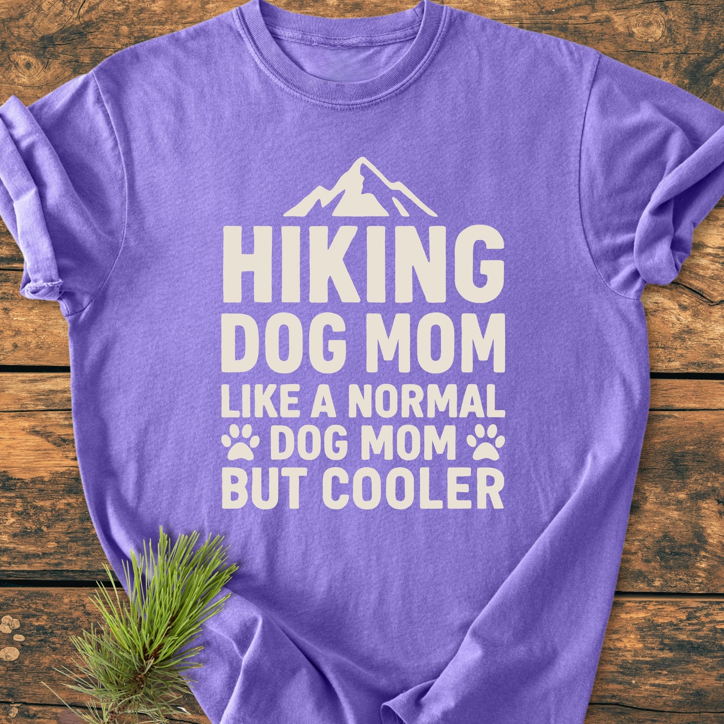 Hiking Dog Mom - Vintage Wash Tee displayed flat on wood, showcasing bold white slogan with mountain and paw icons. Features a pine branch accent, highlighting its rustic and cozy appeal.
