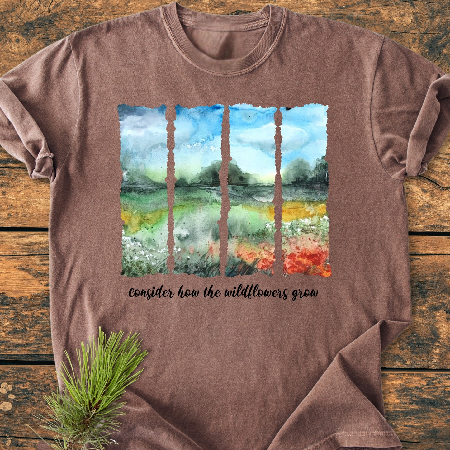 Consider How The Wildflowers Grow - Vintage Heavyweight Tee featuring a watercolor landscape in torn-paper style panels, with script text below. Laid flat on wood, includes a pine sprig detail.