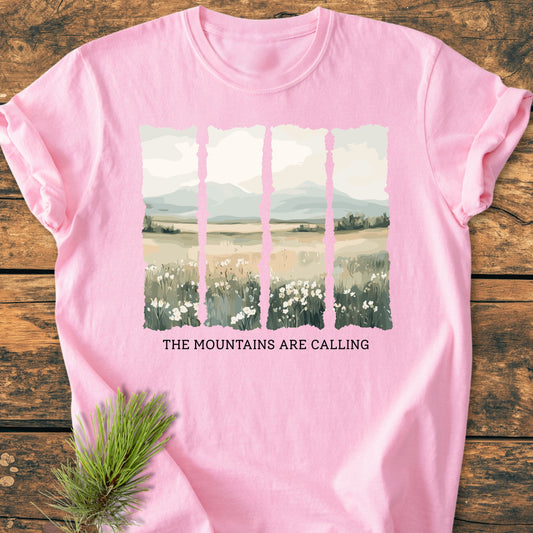 The Mountains Are Calling - Vintage Heavyweight Tee features a misty mountain graphic with wildflowers on a soft pink backdrop, laid flat with a pine bough for a nature-inspired touch.