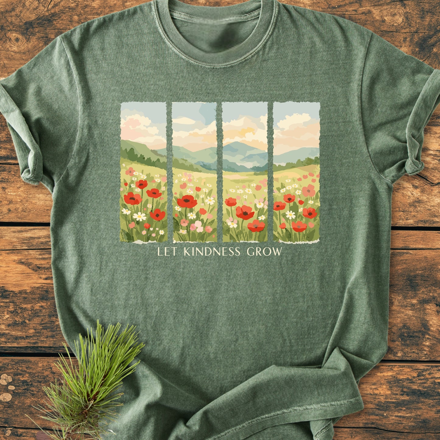 Let Kindness Grow - Vintage Heavyweight Tee featuring a wildflower meadow illustration and slogan on a heather-green background, displayed on rustic wood with a pine sprig beside it.