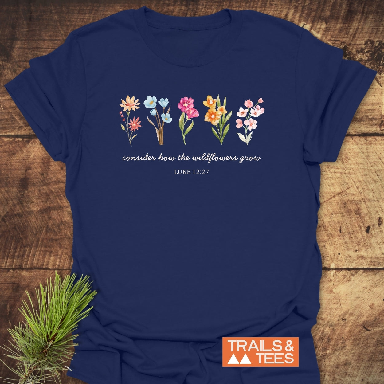 Consider How The Wildflowers Grow, Luke 12:27 T-Shirt displays painted wildflowers with script text on navy fabric, emphasizing outdoor adventure and comfort. Features durable prints, ring-spun cotton, and sustainable production.