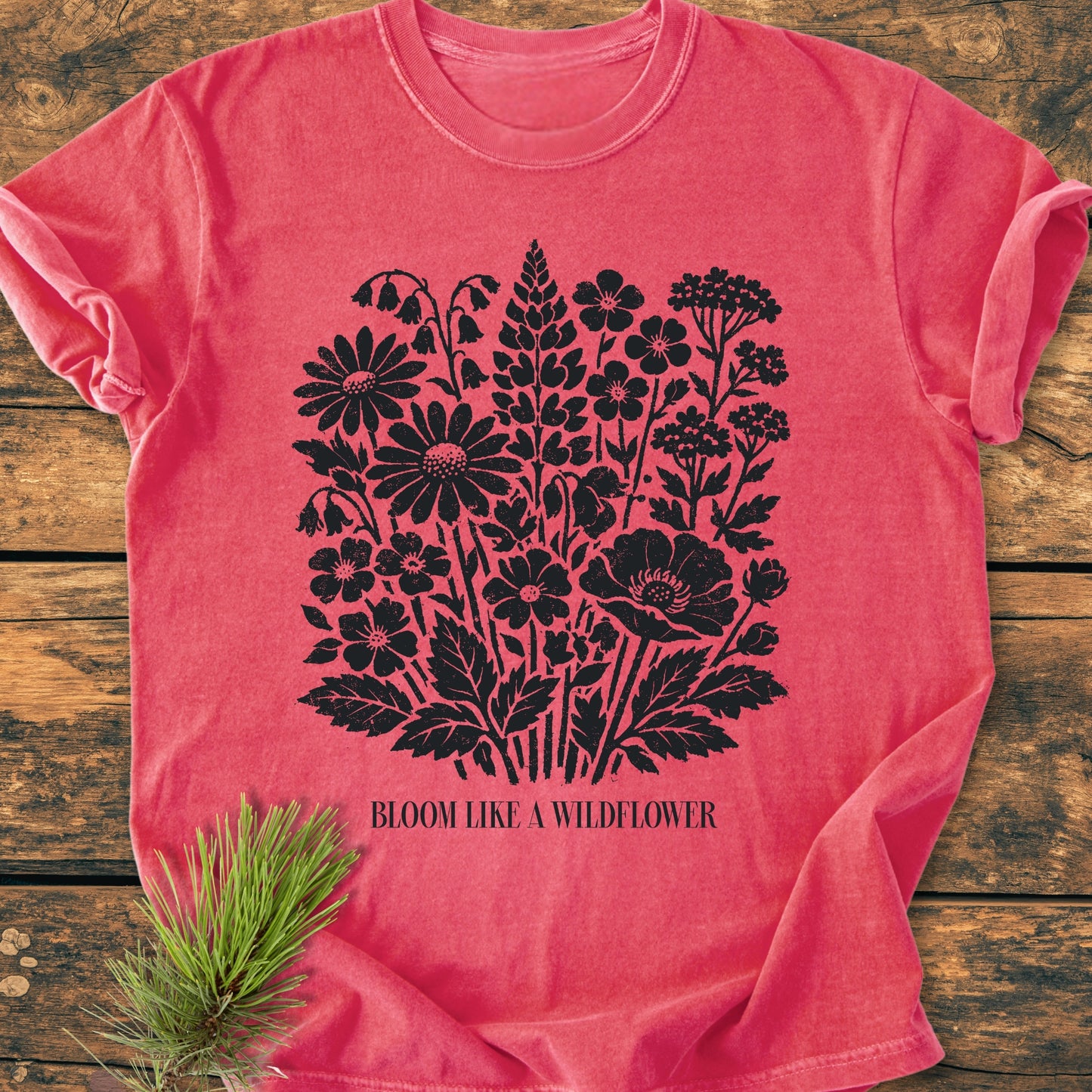 Bloom Like A Wildflower - Vintage Heavyweight Tee, featuring a black wildflower print and slogan, laid flat on a wooden surface with a pine sprig accent.