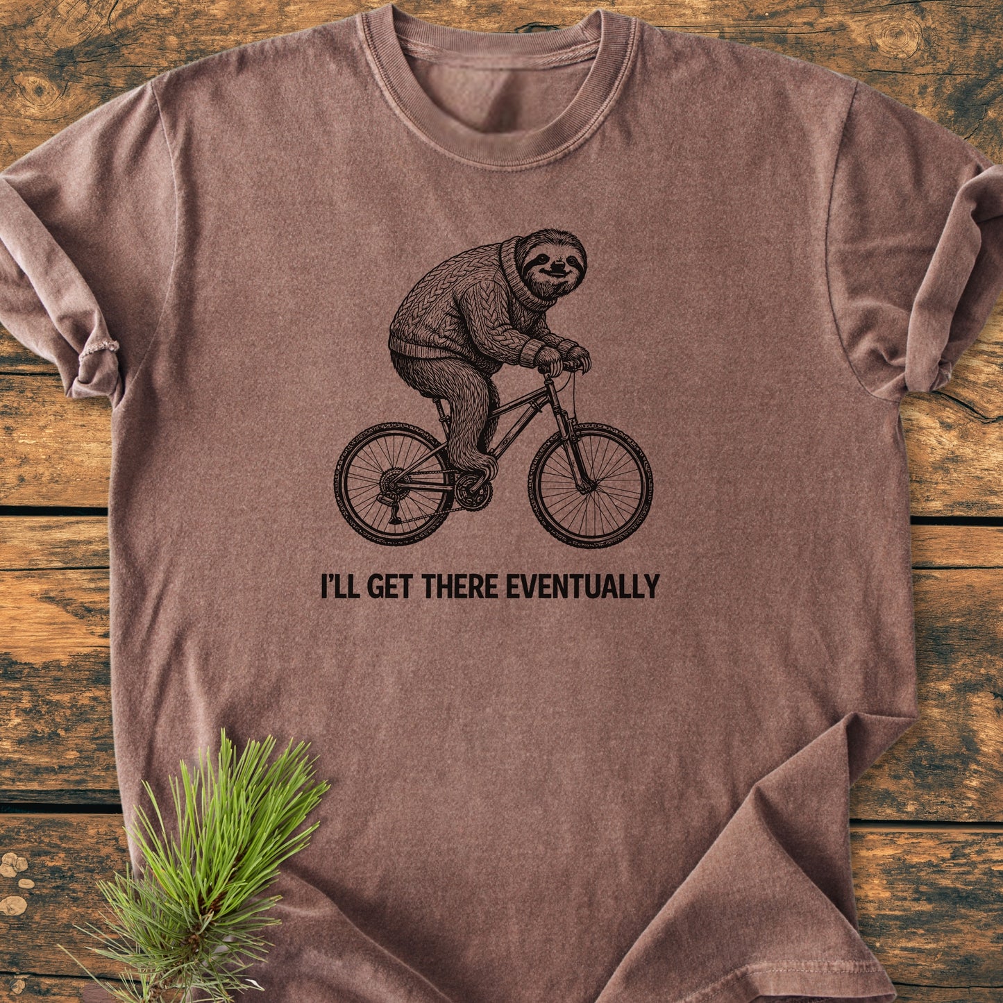 I'll Get There Eventually - Vintage Wash Tee features a sloth in a sweater on a bike, laid flat on a rustic wood background, styled with a pine sprig.