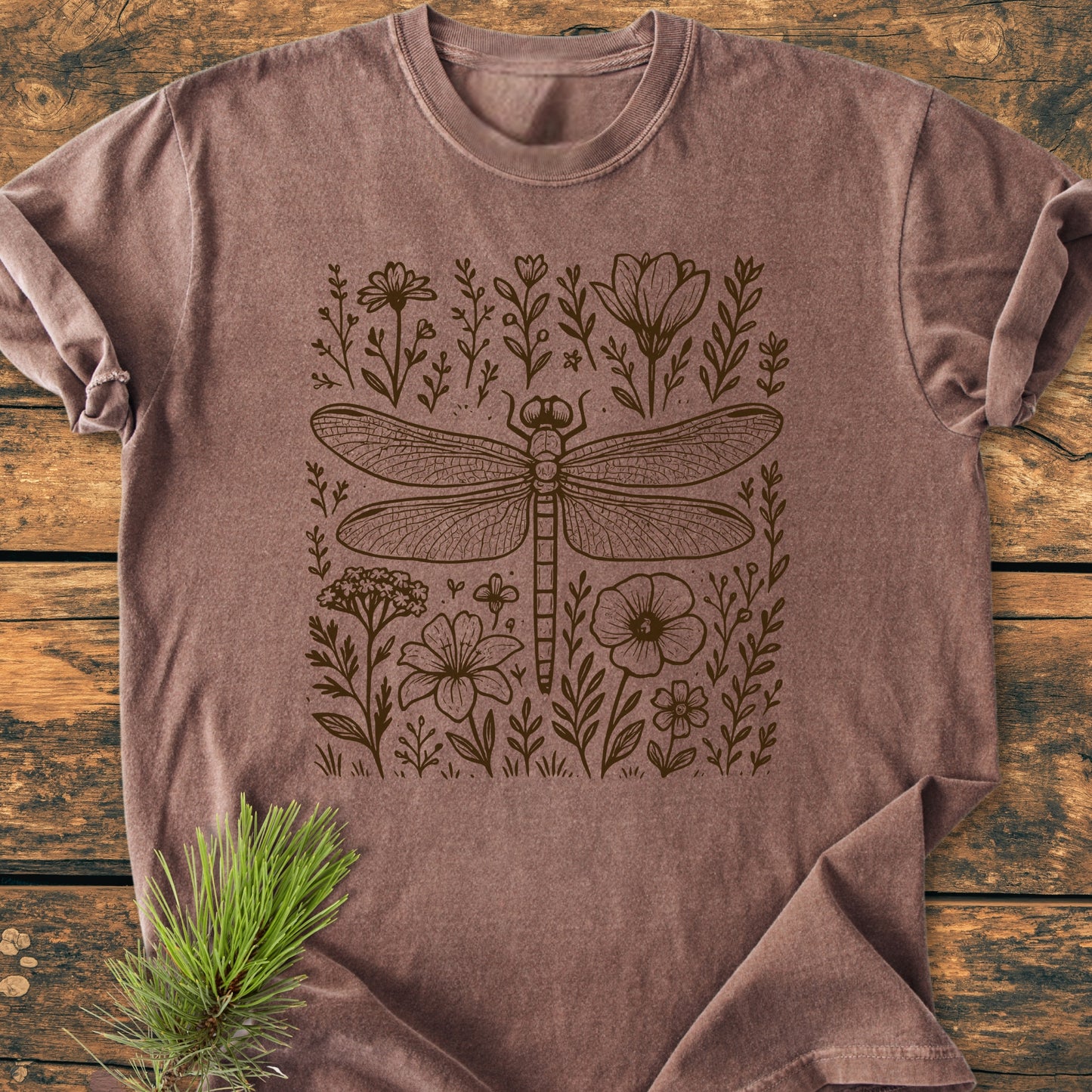 Dragonfly - Vintage Heavyweight Tee features a line-drawing dragonfly with flowers on a heathered brown T-shirt, styled on rustic wood with a pine bough accent, highlighting its relaxed and durable design.