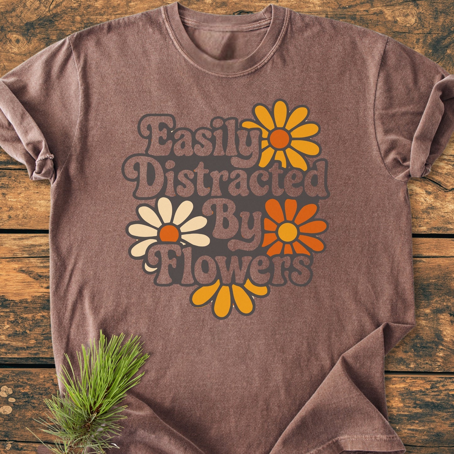 Easily Distracted By Flowers - Vintage Heavyweight Tee, featuring groovy retro font and daisy-like blooms, displayed flat-lay on wood. Pine sprig accentuates the cozy, relaxed-fit Comfort Colors tee.