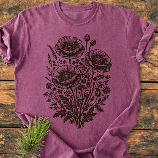 Wildflower Bouquet - Vintage Heavyweight Tee features a black floral design with poppies on a heathered plum T-shirt, displayed on wood. Includes a pine twig accent.