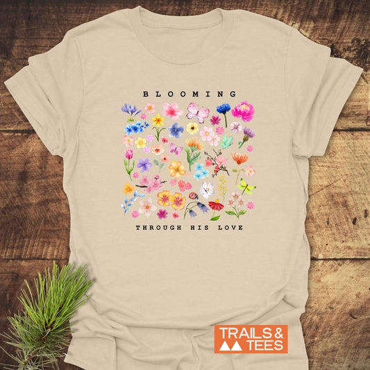 The Blooming Through His Love T-Shirt features a vibrant floral collage with BLOOMING above and THROUGH HIS LOVE below, laid on wood. Ideal for outdoor enthusiasts.