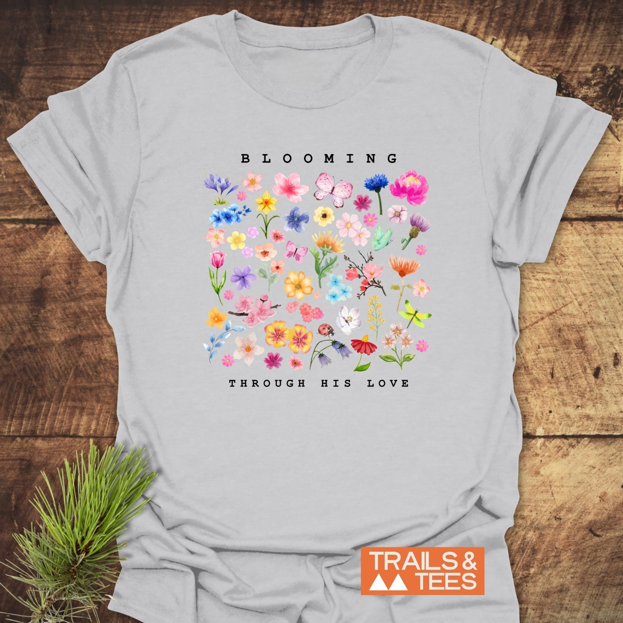 Blooming Through His Love T-Shirt features colorful flower and butterfly illustrations with BLOOMING above and THROUGH HIS LOVE below, plus TRAILS & TEES logo, on soft, lightweight cotton.