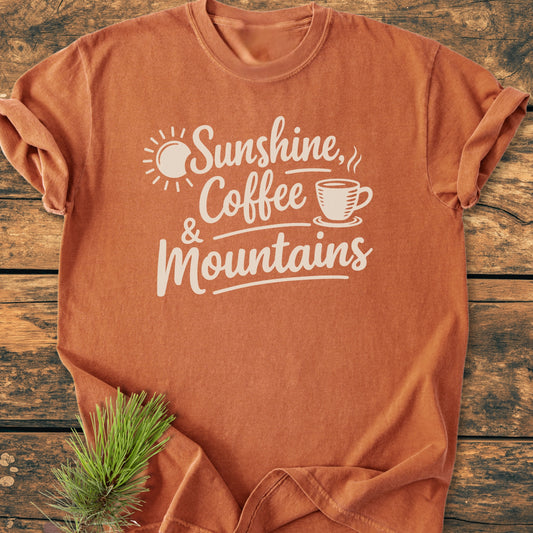 Sunshine, Coffee, Mountains - Vintage Heavyweight Tee featuring a rust-orange hue with white text and graphics. Displayed on wood, with a pine sprig accent. Cozy, relaxed fit, 100% ring-spun cotton.